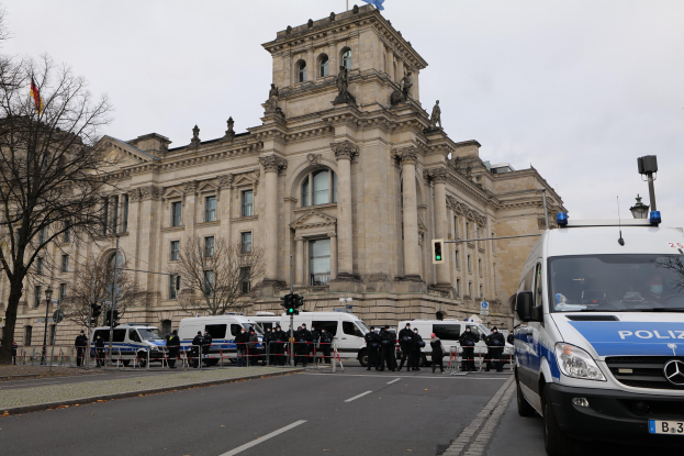 Polizisten vor dem Reichstaggebäude in Berlin, Deutschland, mit Fahrzeugen, einem Zaun, Verkehrsampeln, Laternen, Bäumen und Flaggen im Hintergrund.