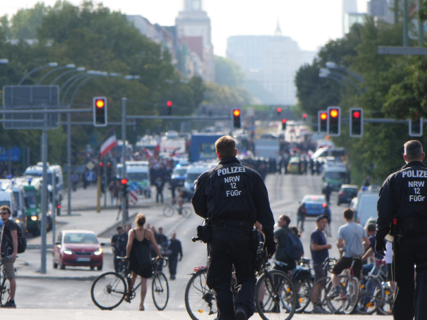 Zwei Polizeibeamte neben einer Gruppe von Radfahrern auf einer Straße mit Fahrzeugen, Verkehrszeichen, Bäumen, Gebäuden und einem blauen Himmel im Hintergrund.