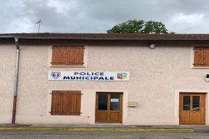Polizeigebäude mit einem Schild, Fenstern, Türen, einer Namenskennzeichnung, einem Rohr, einem Baum und einem bewölkten Himmel im Hintergrund.