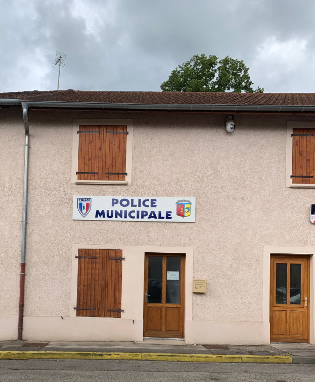 Polizeigebäude mit einem Schild, Fenstern, Türen, einer Namensplakette, einem Rohr, einem Baum und einem bewölkten Himmel im Hintergrund.