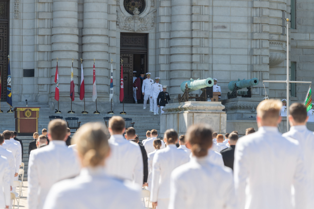 Gruppe von Menschen in weißen Marineuniformen vor einem Gebäude mit Säulen und Treppe, mit Fahnen, einem Podium und Kanonen im Hintergrund.
