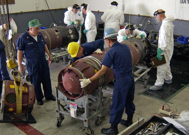 Männer in Helmen und Handschuhen, die eine große Bombe in einer industriellen Fabrikumgebung entschärfen, umgeben von Maschinen und Equipment.