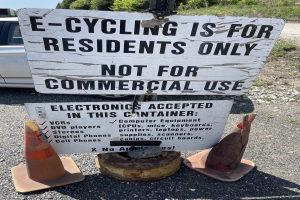 Sign reading "e-cycling is for residents only" placed alongside a road, bordered by traffic cones and other items, with a vehicle, trees, and a clear blue sky in the background.