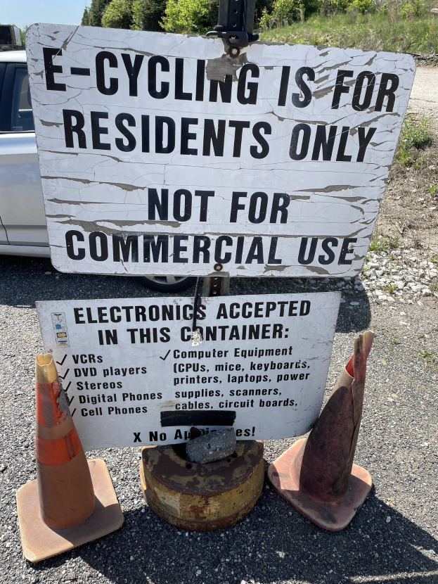Sign reading "e-cycling is for residents only" placed alongside a road, bordered by traffic cones and other items, with a vehicle, trees, and a clear blue sky in the background.