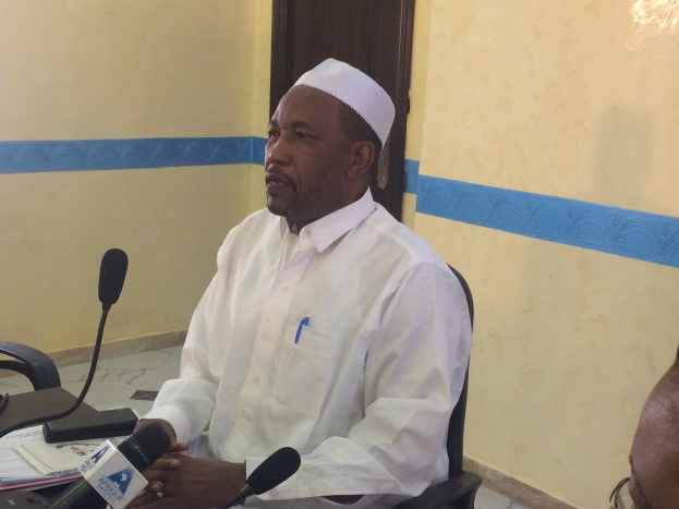 Ein Mann in einem weißen Kleid und Hut sitzt an einem Tisch mit Mikrofonen, spricht, während Papiere und Gegenstände auf dem Tisch sichtbar sind, mit einer anderen Person mit Brille rechts von ihm, in einem Raum mit einer Wand und einer Tür im Hintergrund.