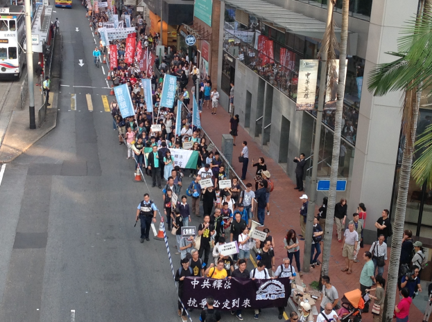 Eine große Gruppe von Menschen marschiert bei einer Demonstration auf einer Stadtstraße, hält Schilder und Plakate.