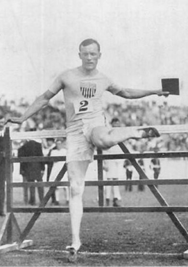 Schwarz-weiß-Foto eines Mannes bei einem Weitsprungwettkampf, mittendrin über ein Hindernis, mit einer nummerierten Shorts und einem T-Shirt, mit Zuschauern im Hintergrund.