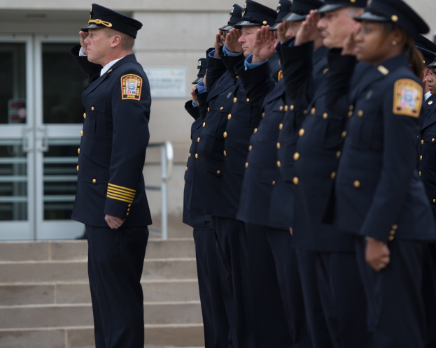 Eine Gruppe von Polizisten in Uniform und mit Mützen, die vor einem Glastür- und Treppenhaus stehend salutieren.