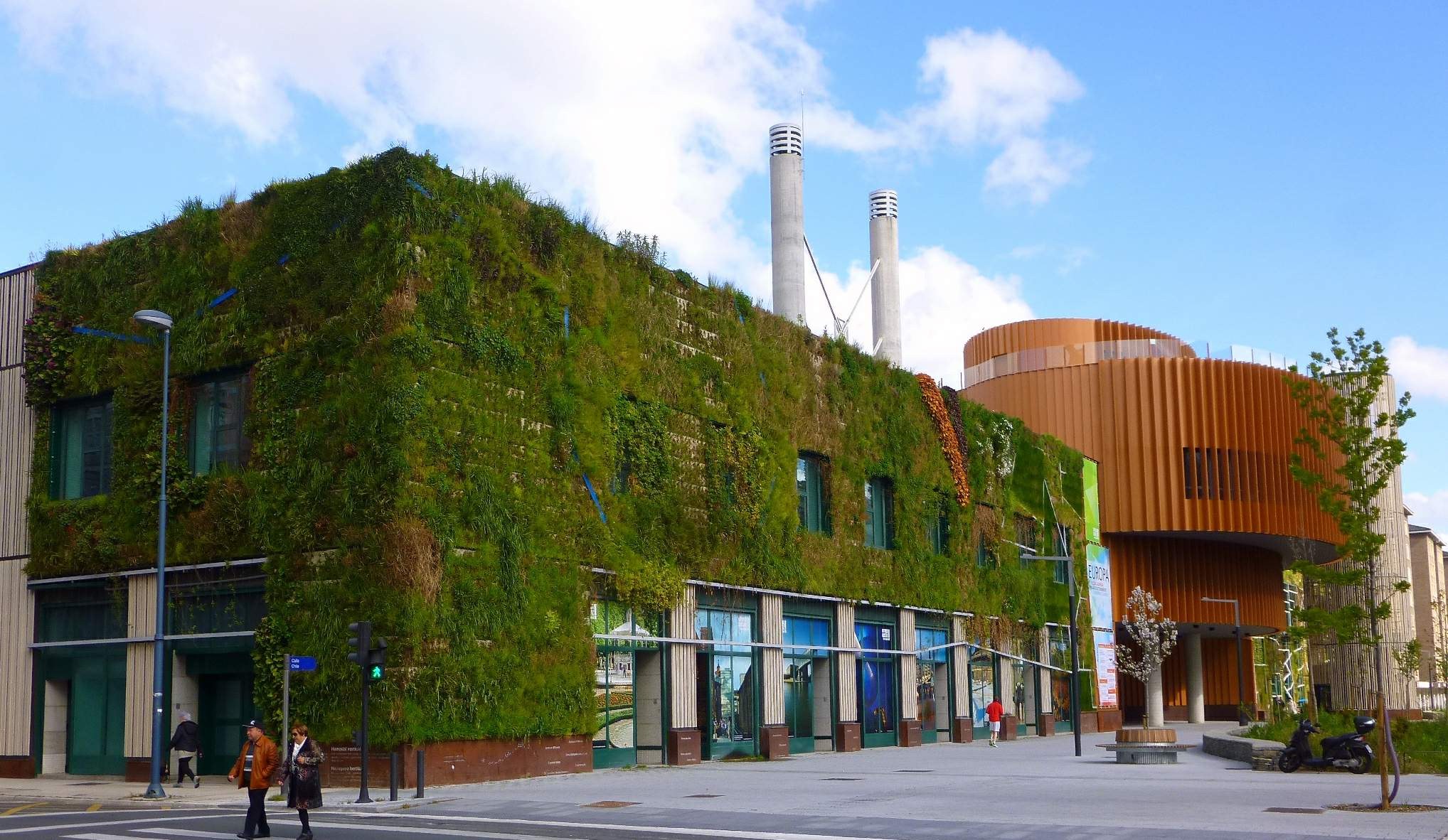 Großes nachhaltiges Gebäude mit einer grünen Wand, umgeben von Straßeninfrastruktur, Fahrzeugen, Fußgängern, Bäumen und einer bewölkten Himmel.