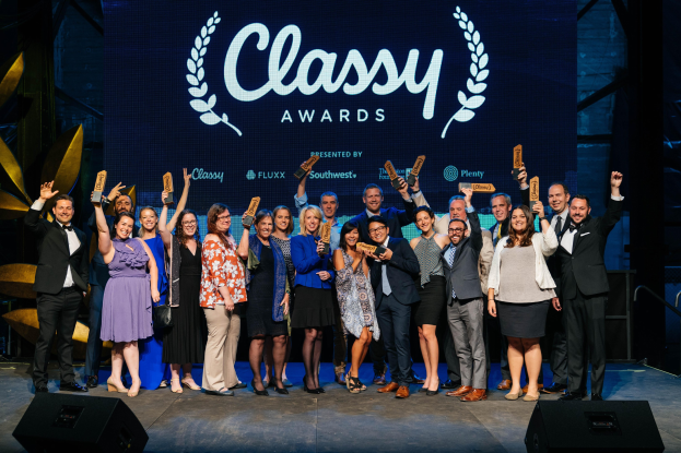 Gruppe von Menschen auf einer Bühne mit Auszeichnungen, mit Lautsprechern darunter und einem Bildschirm im Hintergrund mit der Aufschrift "Classy Awards 2019".