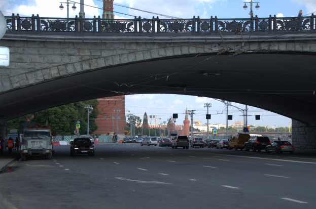 Eine Brücke überspannt eine belebte Straße mit darunterfahrenden Fahrzeugen; Fußgänger stehen auf dem Gehweg; Gebäude, Bäume, Laternen, Verkehrssignale und der Himmel sind im Hintergrund zu sehen.