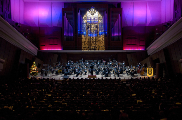 Großer Saal mit Publikum bei einem Konzert; Musiker auf der Bühne spielen Instrumente und andere sitzen in Stühlen; Weihnachtsbaum und geschmückte Wände mit Lichtern im Hintergrund.