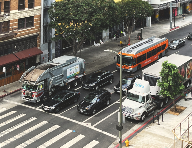 Eine belebte Stadtstraße mit verschiedenen Fahrzeugen, Fußgängern, Gebäuden, Bäumen und Straßeneinrichtungsgegenständen.