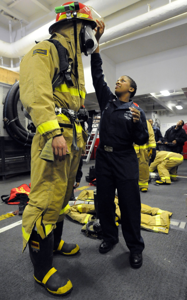 Zwei Feuerwehrleute, eine in einem schwarzen Kleid mit Helm und ein anderer in einer gelben und schwarzen Uniform, stehen zusammen in einer Gruppe von Menschen und Feuerlöschausrüstung.