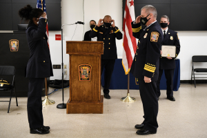 Gruppe uniformierter Personen in Masken vor einem Podium mit Mikrofon, umgeben von Fahnen, Stühlen und Tischen, während einer Vereidungszeremonie für einen neuen Feuerwehrchef.