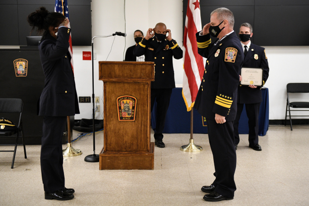Gruppe uniformierter Personen in Masken vor einem Podium mit Mikrofon, umgeben von Fahnen, Stühlen und Tischen, während einer Vereidungszeremonie für einen neuen Feuerwehrchef.