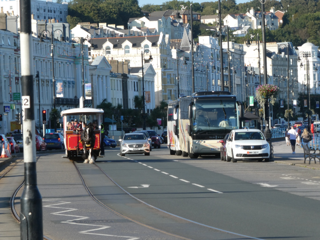 Eine belebte Stadtstraße mit hohen Gebäuden, Fahrzeugen, Fußgängern, Ampeln, Laternenpfählen, Bäumen und einem klaren blauen Himmel.