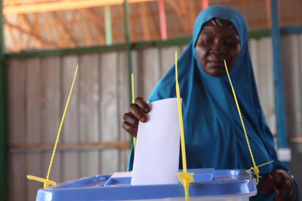 Frau mit blauem Kopftuch hält ein Blatt Papier in der Hand und steht neben einer Kiste mit Stöcken, wahrscheinlich beim Abstimmen an einer Wahllokal mit einer Wand und einem Schuppen im Hintergrund.