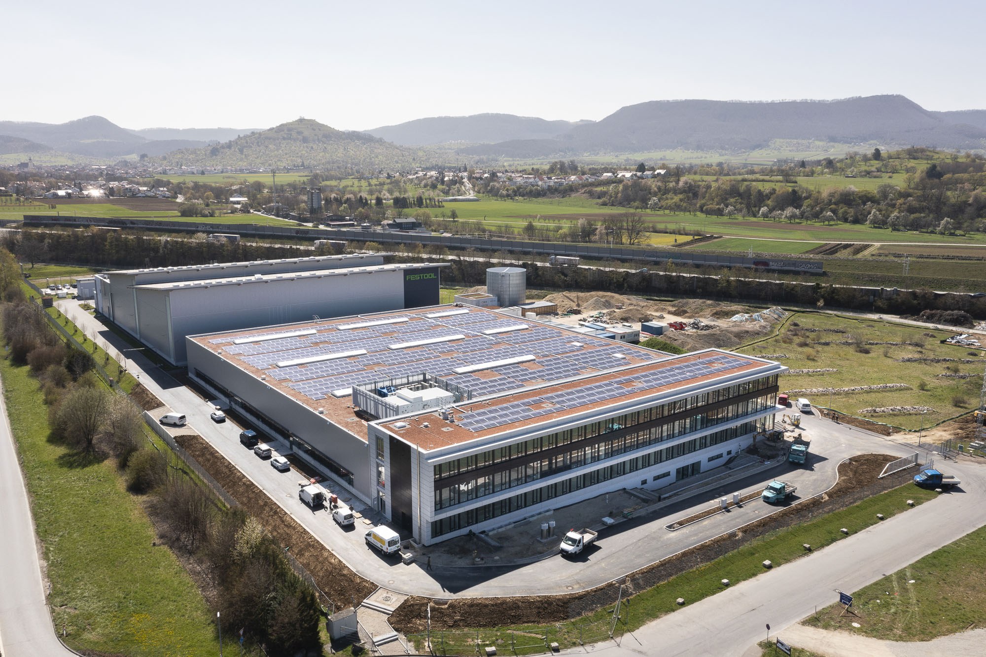 Luftaufnahme eines großen Fabrikgebäudes mit Solarmodulen auf dem Dach, umgeben von Fahrzeugen, Bäumen, Gras und Strommasten, mit Hügeln und einem klaren blauen Himmel im Hintergrund.