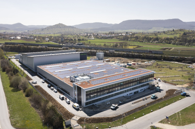 Luftaufnahme eines großen Fabrikgebäudes mit Solarmodulen auf dem Dach, umgeben von Fahrzeugen, Bäumen, Gras und Strommasten, mit Hügeln und einem klaren blauen Himmel im Hintergrund.