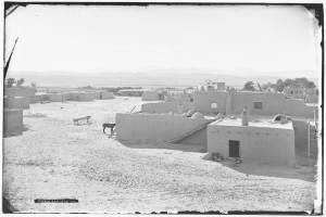Schwarzes und weißes Foto einer Adobe-Dorfschaft in einer Wüstenlandschaft mit Häusern, Bäumen, Bergen im Hintergrund, Tieren am Boden und Himmel darüber.
