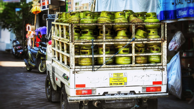 Ein Lastwagen mit Gasflaschen beladen, der am Straßenrand geparkt ist, mit Gebäuden, Bäumen und einem Geschäft im Hintergrund.