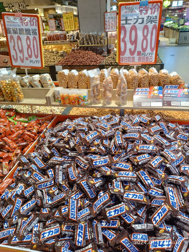 Vitrine voller verschiedener Süßigkeiten, darunter Snickers-Riegel, in einem Geschäft mit Lebensmitteln auf Regalen und Hintergrunddetails.