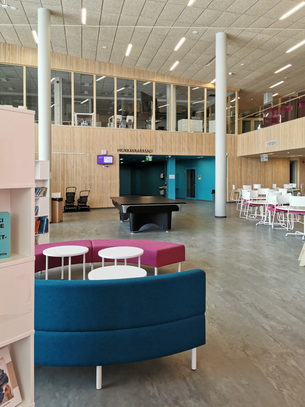 Ein geräumiger Raum, der einer Schulbibliothek ähnelt, mit Tischen, Stühlen, Sofas, Bücherregalen, Säulen, Deckenleuchten und einem Wandfernseher.