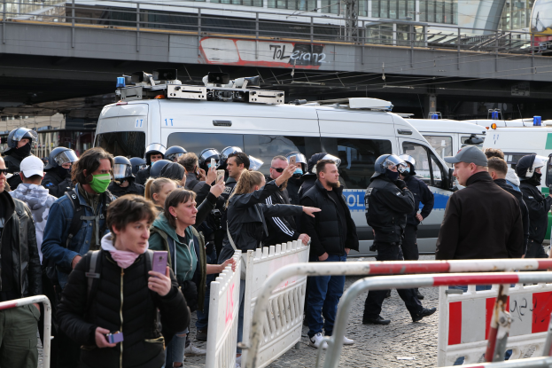 Eine Gruppe von Menschen steht vor Polizeifahrzeugen mit Barrieren im Vordergrund und einer Brücke im Hintergrund während einer Demonstration in Berlin, Deutschland.