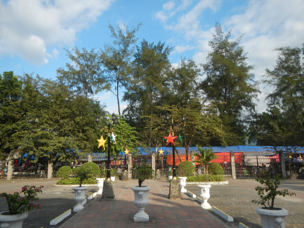 Ein Park mit Topfpflanzen und Bäumen in der Mitte, umgeben von Straßenmasten, Straßenlaternen, Dekorationsgegenständen und Grills unter einem bewölkten Himmel.