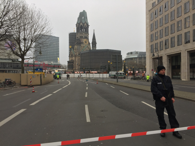 Ein Polizeibeamter in schwarzer Uniform und Mütze steht in einer Berliner Straße umgeben von Absperrbaken, Fahrrädern, Laternen, Bäumen und Gebäuden, im Hintergrund ist ein Kirchturm und ein klarer blauer Himmel zu sehen.