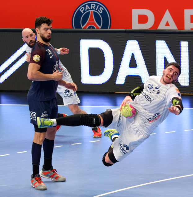 Eine Gruppe von Männern, die auf einem Handballfeld spielen, wobei einer einen Ball hält; ein Paris Saint-Germain-Futsal-Siegertafel ist im Hintergrund zu sehen.