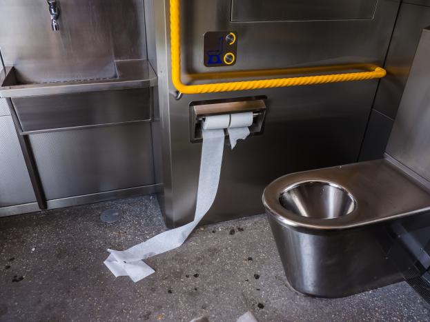 Stahltoilette mit einer Rolle Toilettenpapier daneben, ein Waschbecken mit Wasserhahn und verschiedene Gegenstände auf dem Boden.
