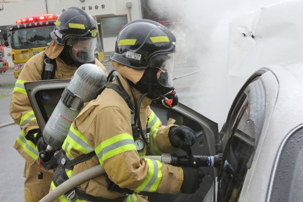 Zwei Feuerwehrleute in Schutzausrüstung verwenden einen Schlauch, um ein brennendes Auto zu löschen, wobei Rauch aufsteigt und Fahrzeuge sowie ein Gebäude im Hintergrund zu sehen sind.