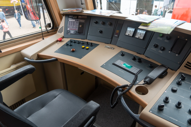 Leitstelle eines Zuges mit einem Schreibtisch, Stühlen, elektronischen Geräten, Papieren und durch Glasfenster sichtbaren Menschen draußen.