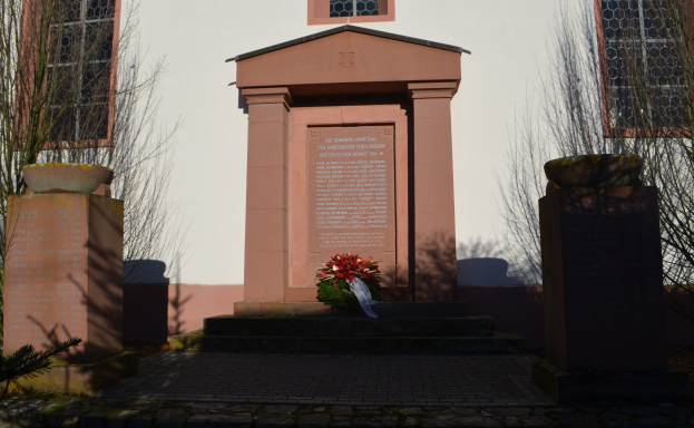 Ein Gebäude mit Fenstern, ein Denkmal mit Text, ein Blumenbouquet auf dem Boden, Topfpflanzen, eine Baumgruppe und ein bewölkter Himmel, mit einem Grab und einem Kranz davor.