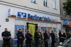 Eine Gruppe von Polizeibeamten vor einer Berliner Volksbank, einige halten Helme, mit einem Auto auf der rechten Seite und einem Baum auf der linken Seite; Gebäude mit Fenstern und Texttafeln im Hintergrund.