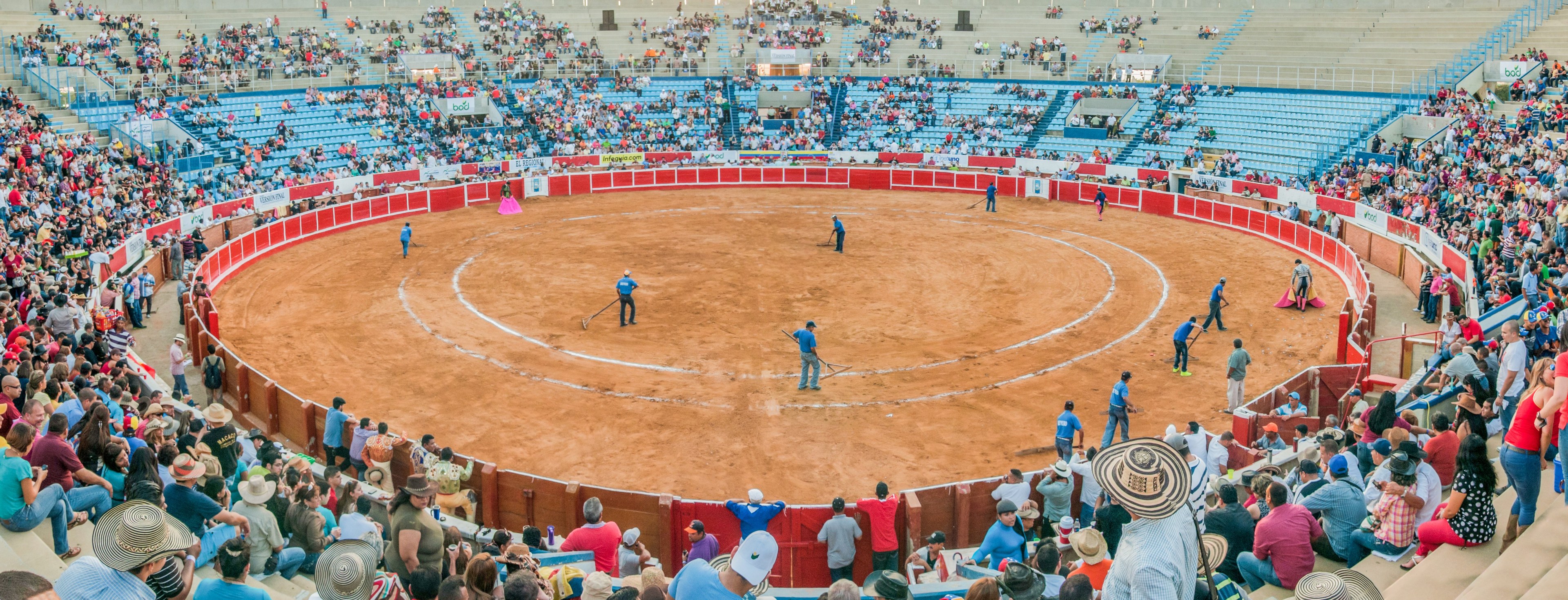 Eine große Menschenmenge schaut bei einer Stierkampfveranstaltung in einem Stadion zu, wobei einige stehen, sitzen oder Gegenstände halten, und eine Person mit Hut unten.