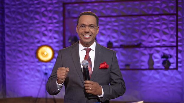 Stephen Colbert, Moderator von *The Daily Show with Trevor Noah*, steht auf einer Bühne und hält ein Mikrofon in der Hand, trägt einen Anzug, eine Krawatte und eine Brille und hat ein Licht und einen Ständer hinter sich.