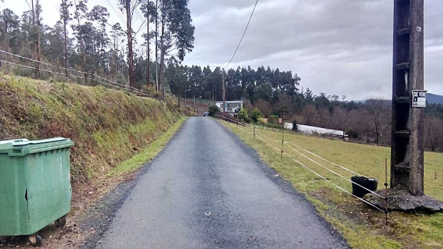 Ein grüner Müllcontainer sitzt neben einer Straße, die mit Gras, Pflanzen, Bäumen, Masten, Drähten und einem Zaun gesäumt ist, unter einem bewölkten Himmel, mit einem Wegweiser auf der rechten Seite und verstreuten Gegenständen auf der linken Seite.