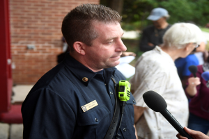 Ein uniformierter Polizist spricht vor Reportern im Freien, hält ein Mikrofon und ist von anderen umgeben, die ebenfalls Mikrofone halten.