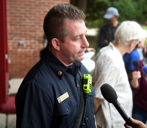 Ein uniformierter Polizist spricht vor Reportern im Freien, hält ein Mikrofon und ist von anderen umgeben, die ebenfalls Mikrofone halten.