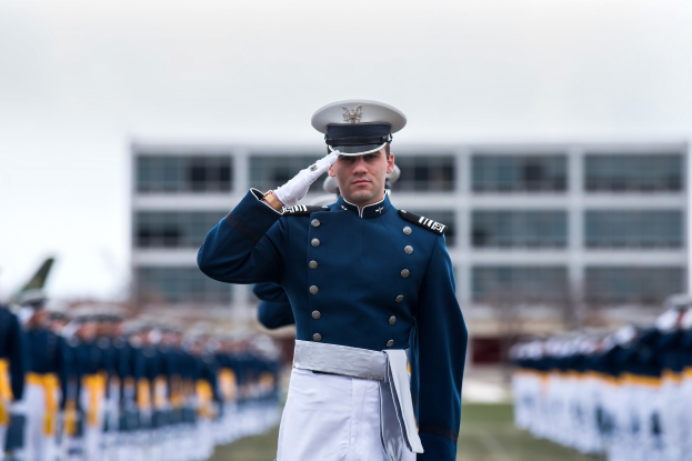 Ein Mann in militärischer Uniform salutiert auf einer Abschlussfeier, umgeben von einer Menge, mit einem Gebäude und Himmel im Hintergrund.
