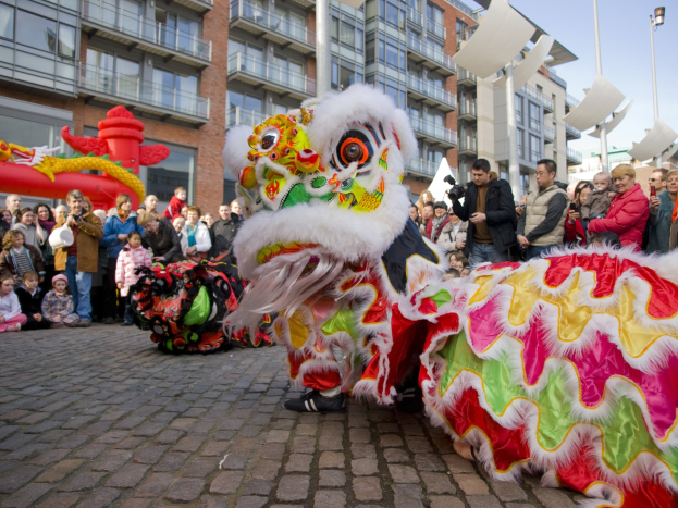 Ein lebendiges chinesisches Neujahrsfest in Amsterdam mit einem Löwen Tanz und einer Menge Schaulustiger, einige fotografieren das Ereignis, vor Buildings und einem klaren blauen Himmel.