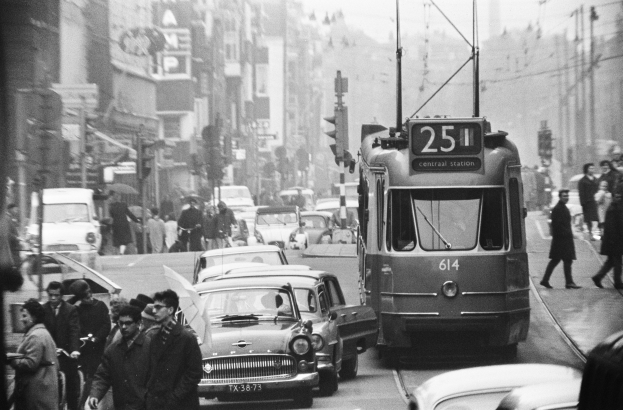 Schwarze und weiße Stadtstraße mit starkem Verkehr, Fußgänger auf Gehwegen mit Regenschirmen und Gebäuden im Hintergrund.