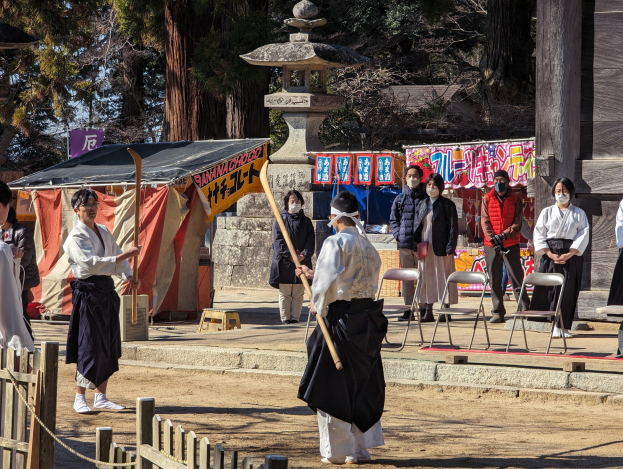 Eine Gruppe von Menschen in traditioneller Kleidung versammelt sich im Freien in Kyoto, einige tragen Masken und halten Holzstöcke, mit Bannern, Stühlen, einem Zelt und einem Holzzaun gegen einen klaren blauen Himmel.