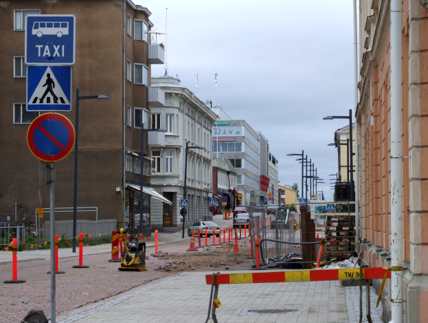 Stadtstraße mit Gebäuden, Straßeninfrastruktur, Fahrzeugen, einer Baustelle, Bäumen und einem bewölkten Himmel.
