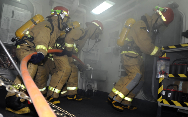 Feuerwehrleute in Schutzausrüstung mit Helmen und Sauerstoffflaschen arbeiten an einem Löschfahrzeug, mit einem Gestell voller Gegenstände und Hintergrundrohren und Lichtern.