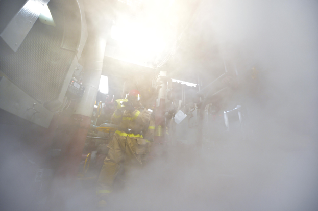 Feuerwehrleute in Schutzausrüstung arbeiten in einem raucherfüllten Raum mit verstreuten Gegenständen.