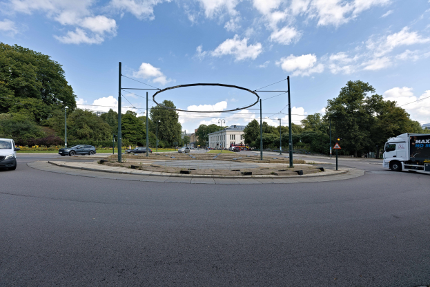 Kreisverkehr mit Fahrzeugen, die darum fahren, umgeben von Pfählen, Bäumen, Gebäuden und einem bewölkten Himmel.